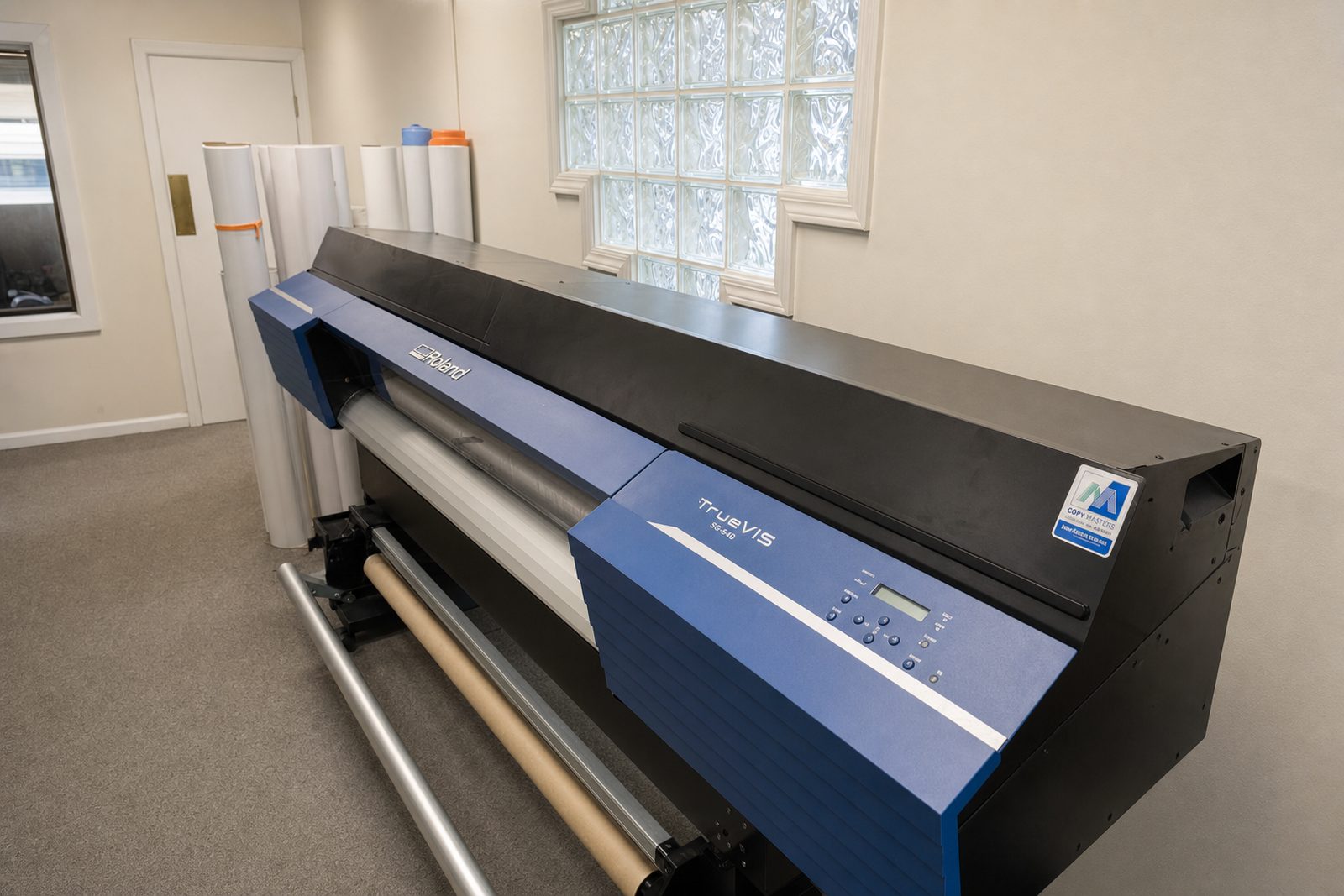 The shop's Roland TrueVIS wide-format printer, with rolls of banner stock standing by the glass-block window